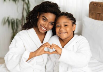 Happy Black Mom And Daughter In Bathrobes Making Heart Sign Form Hands, Connecting Their Arms, Having Fun And Bonding At Home, Relaxing On Bed Together After Bath, Smiling At Camera, Closeup
